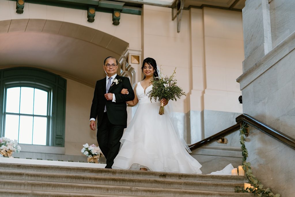 The bride and her father walk down the decorated staircase of Joliet Union Station framed by greenery and flowers.