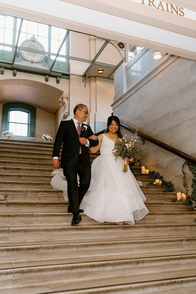 The bride and her father walk down the decorated staircase of Joliet Union Station framed by greenery and lit candles.