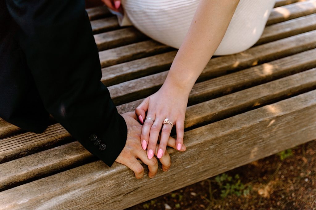 A bride holds the hand of her future groom while sitting on a bench.  You can see her engagement ring.  