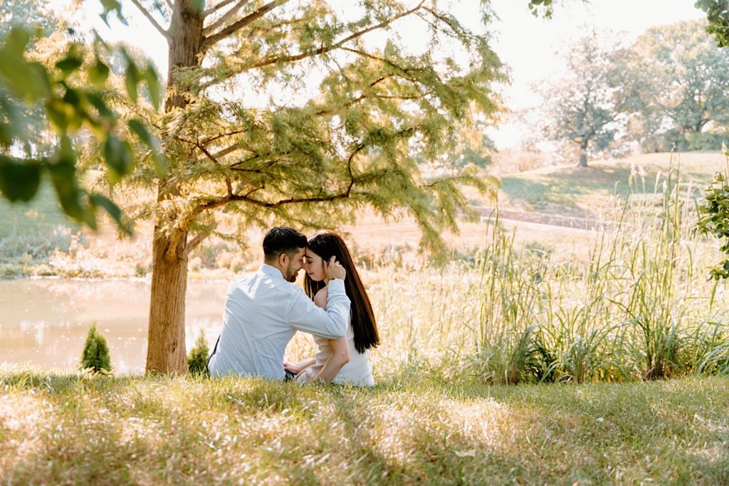 Fiancé's sit together on a grass covered hill overlooking a lake at the Chicago Botanic Gardens.