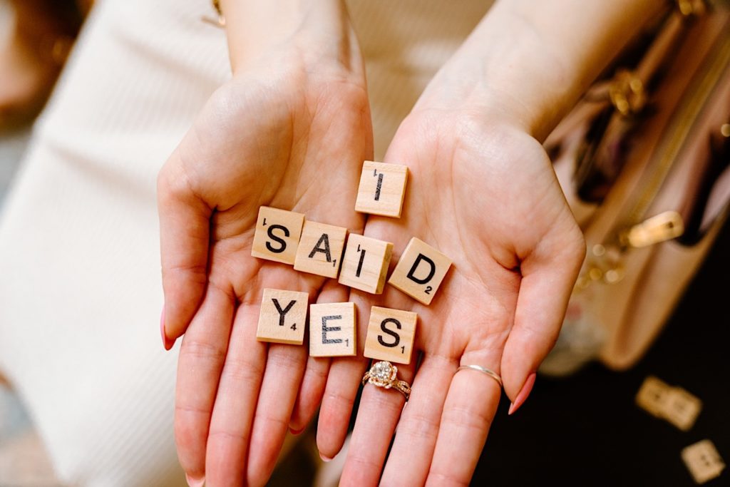 A fiancée holds wooden scrabble letters that say I Said Yes with her ring on backwards facing the camera.
