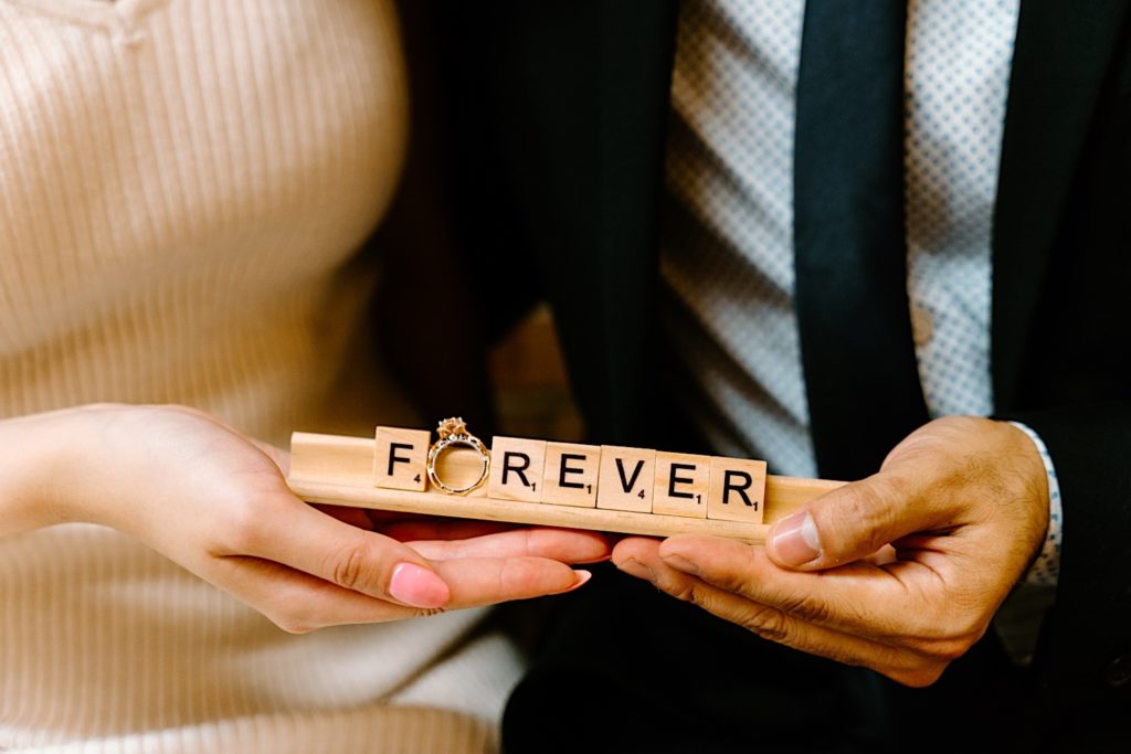 Fiancés hold a wooden scrabble holder with the word Forever in scrabble pieces across it and her engagement ring taking the place of the o.