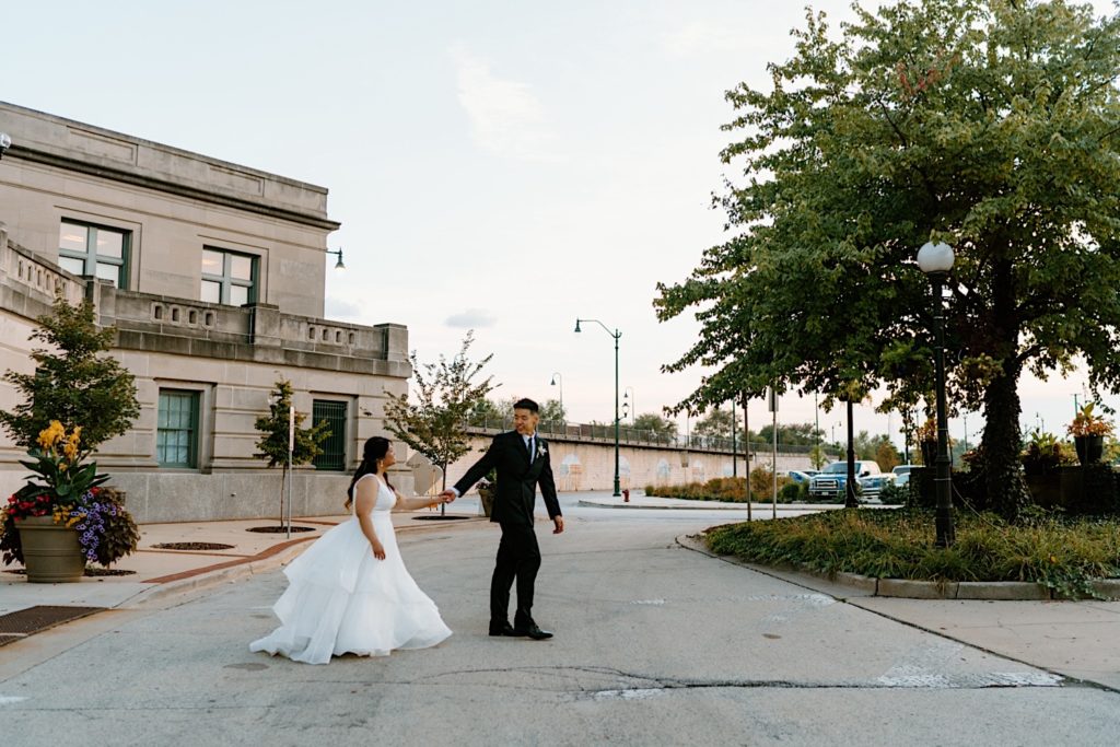 A bride and groom hold hands and walk across the street in the parking lot at Joliet Union Station.