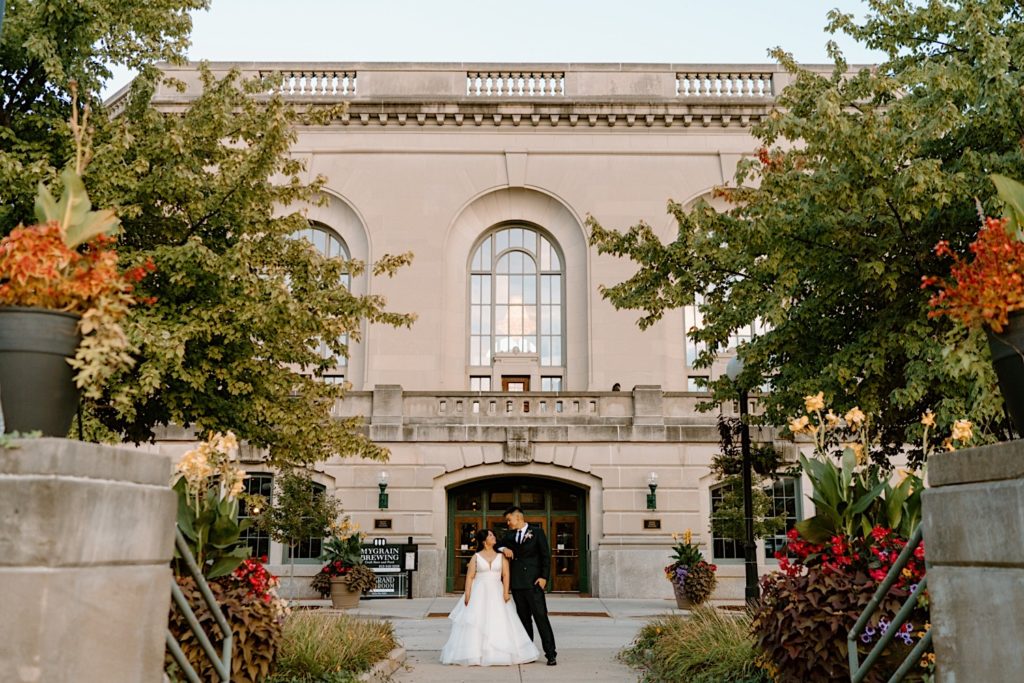 A bride and groom stand in the gardens outside of Joliet Union Station during their wedding portraits holding hands and looking at one another.