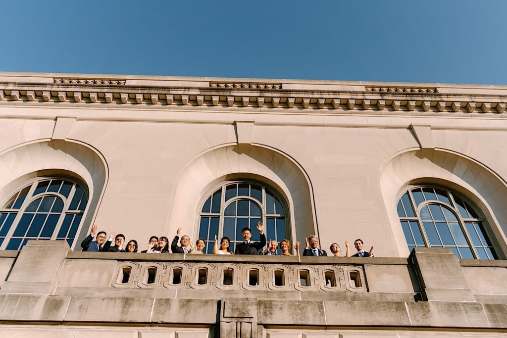 A wedding party stands looking over the balcony smiling and waving at the camera outside of Joliet Union Station.