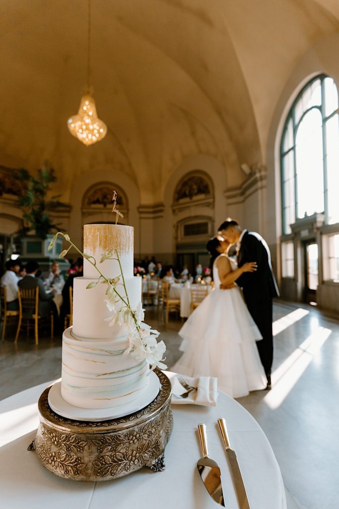 A bride and groom stand kissing with their wedding cake in the foreground. 