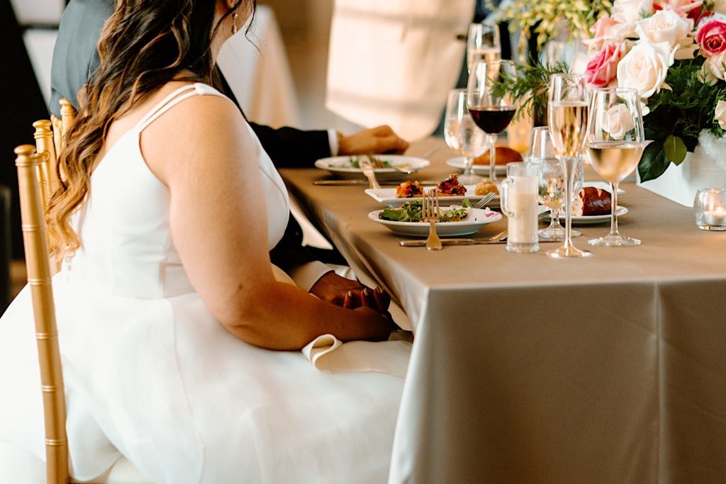 A bride and groom hold hands underneath the table during their dinner.