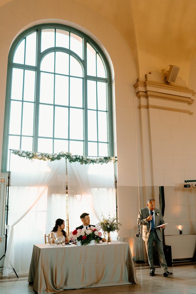 A bride and groom watch as one of their guests toasts to them during their wedding reception.
