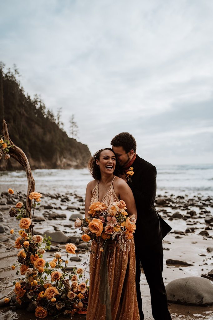 A bride and groom smile at the camera standing on a beach in Oregon during their elopement.