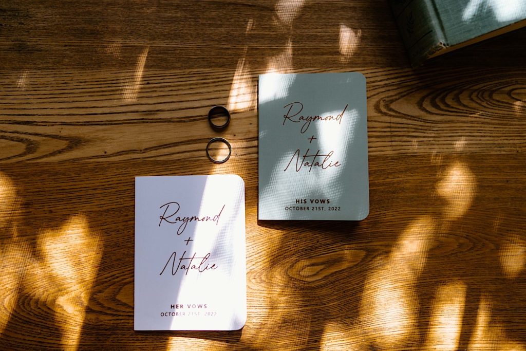 A bride and grooms vow books underneath the shimmering shadows of trees on a wood table.