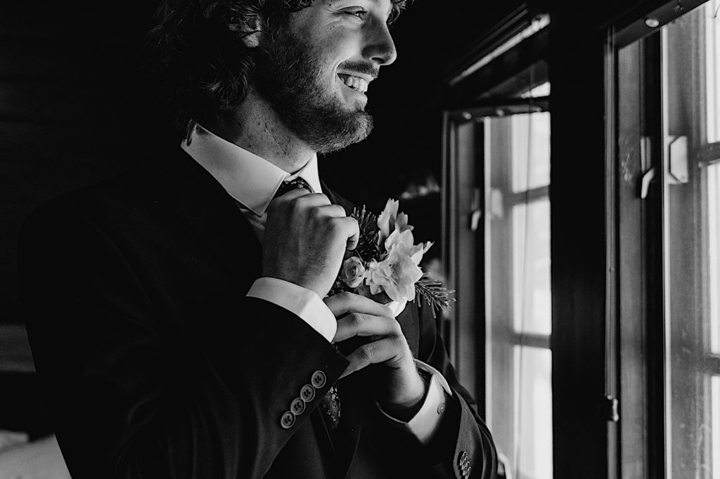 A groom stands looking out the window while tightening his tie smiling. 