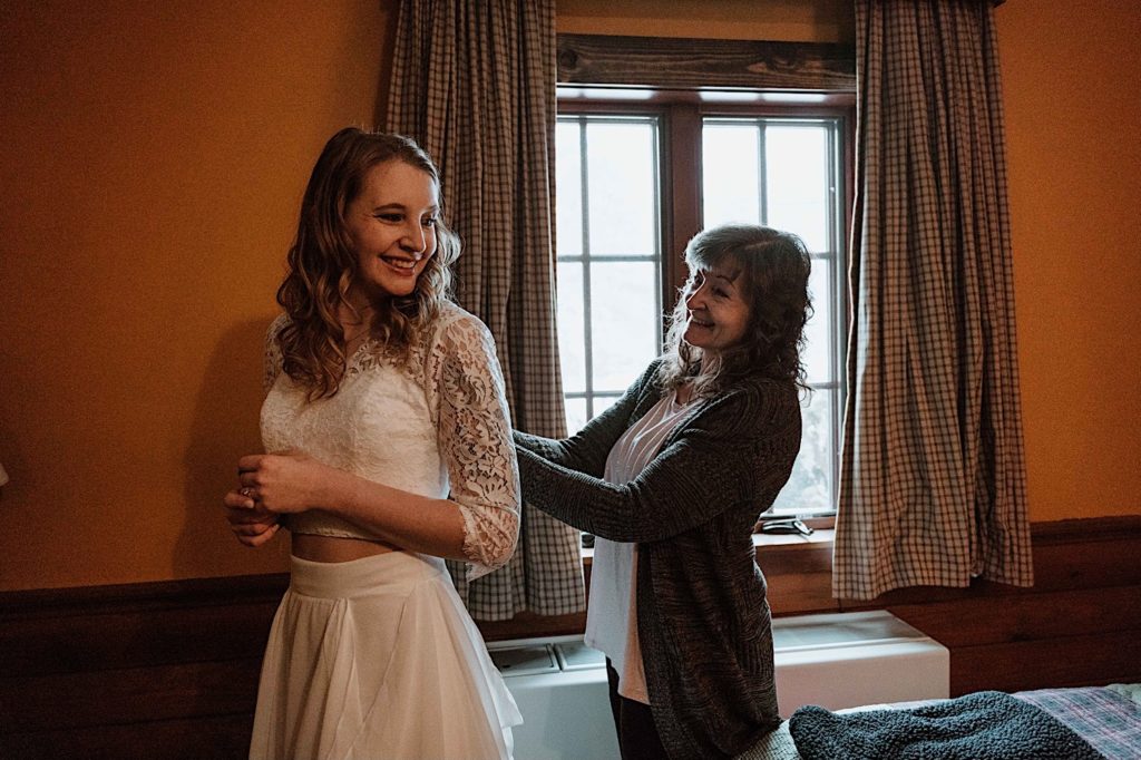 A bride smiles while her mom buttons the back of her two piece wedding dress before her elopement at Starved Rock State Park.