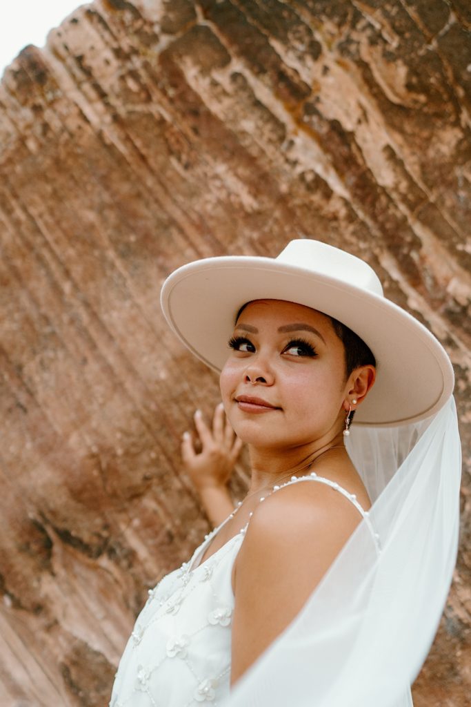 A bride stands in front of Red Rocks looking back at her groom.  She wears a wide brim hat with a veil.