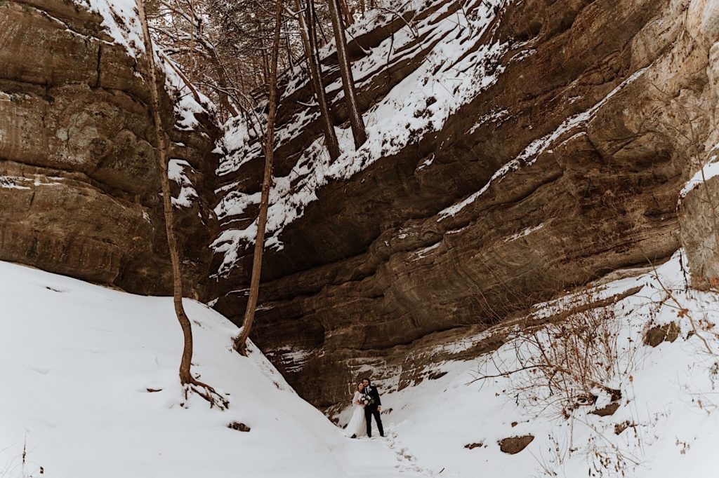 A bride and groom stand in the snow next to a cliff while smiling at the camera.
