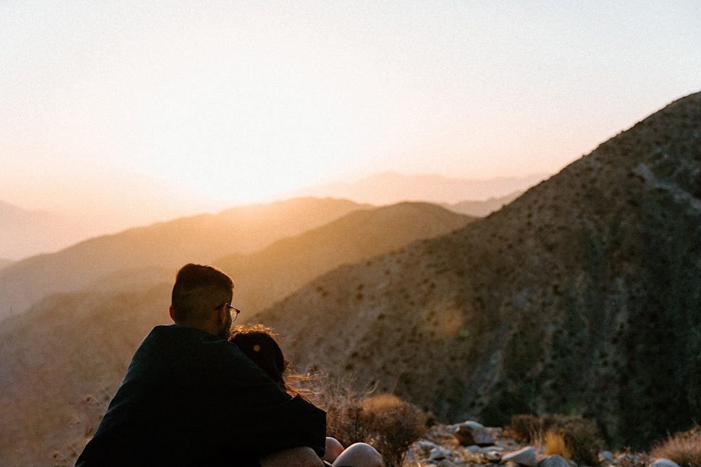 A bride and groom hold one another while watching the sun set over the desert.