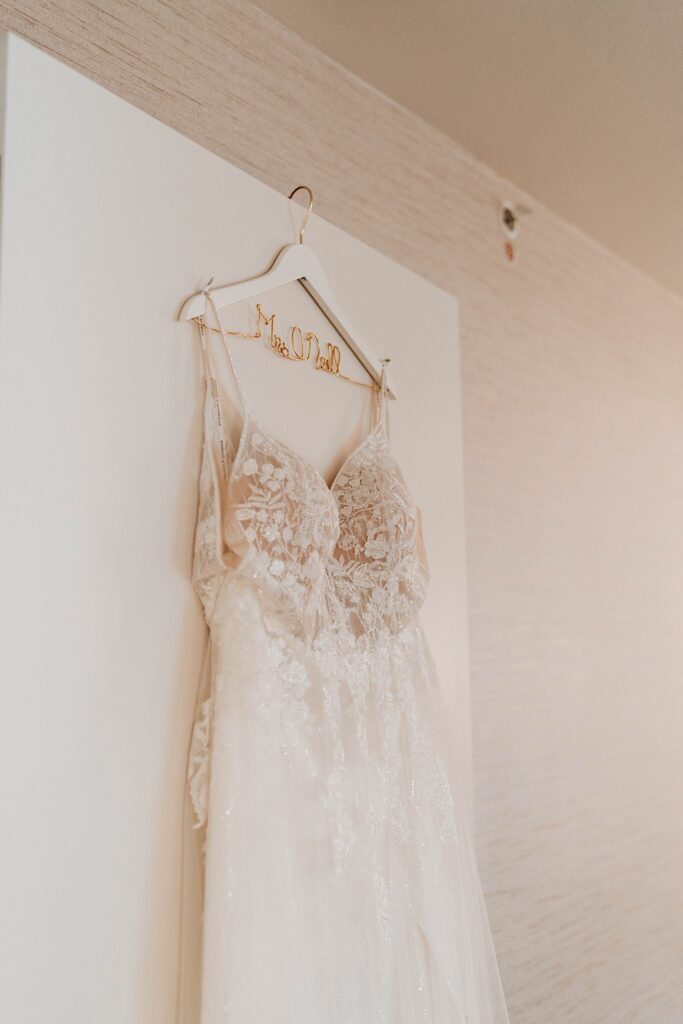 A lace wedding dress hangs from a hotel room door hanging from a white hanger with the brides future last name scripted in the wire.  