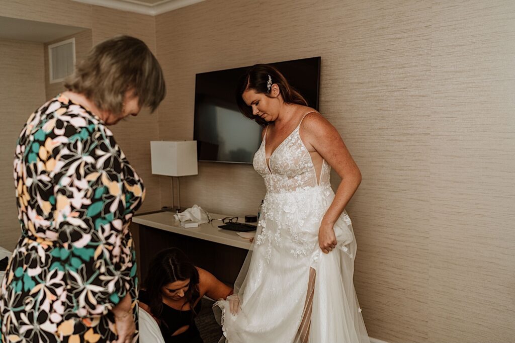 Bride holds her dress up while her bridesmaid puts on her shoes in their hotel room before their wedding.