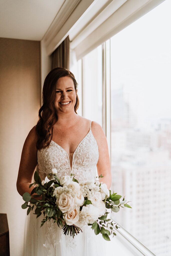 A bride holds her bouquet of white roses standing next to a window in her Chicago hotel before her wedding at the 95th floor Signature Room.