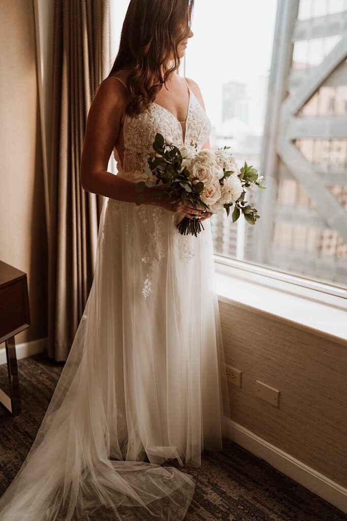 A bride holds her bouquet of white roses standing next to a window in her Chicago hotel before her wedding at the 95th floor Signature Room.