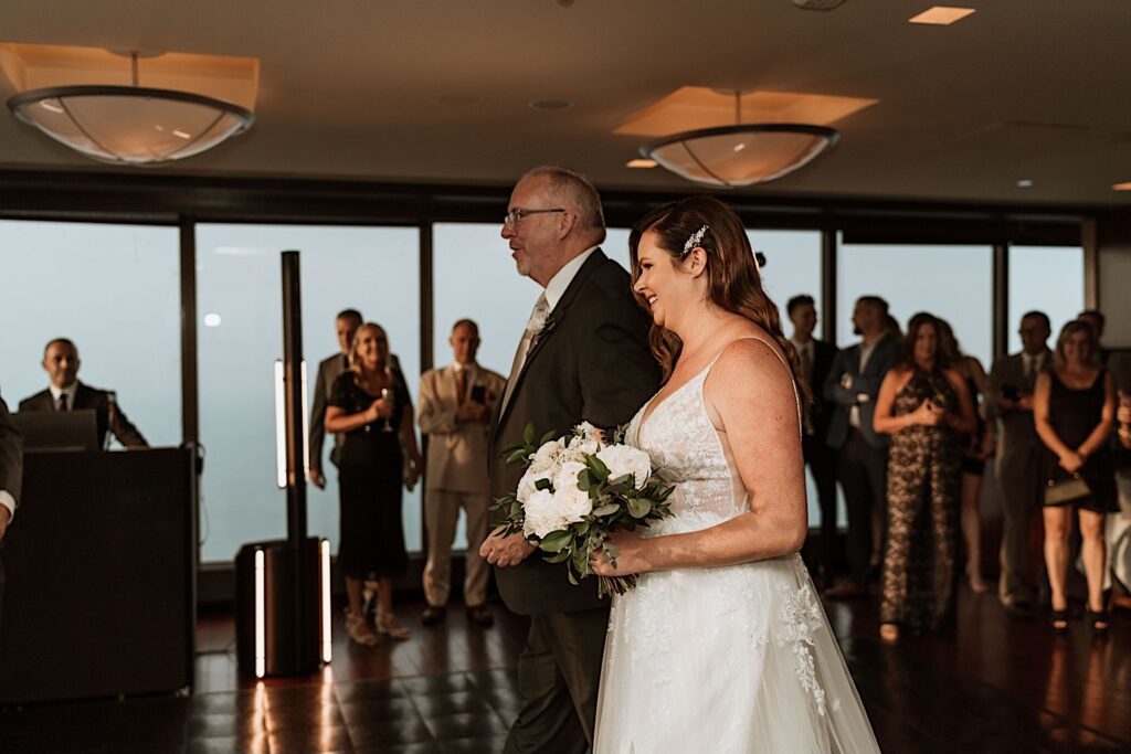 A bride holds her fathers arm while walking to her groom during their ceremony on the 95th floor.