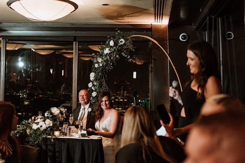 A bride and groom smile and laugh at a speech given by a guest during their wedding reception on the 95th floor.