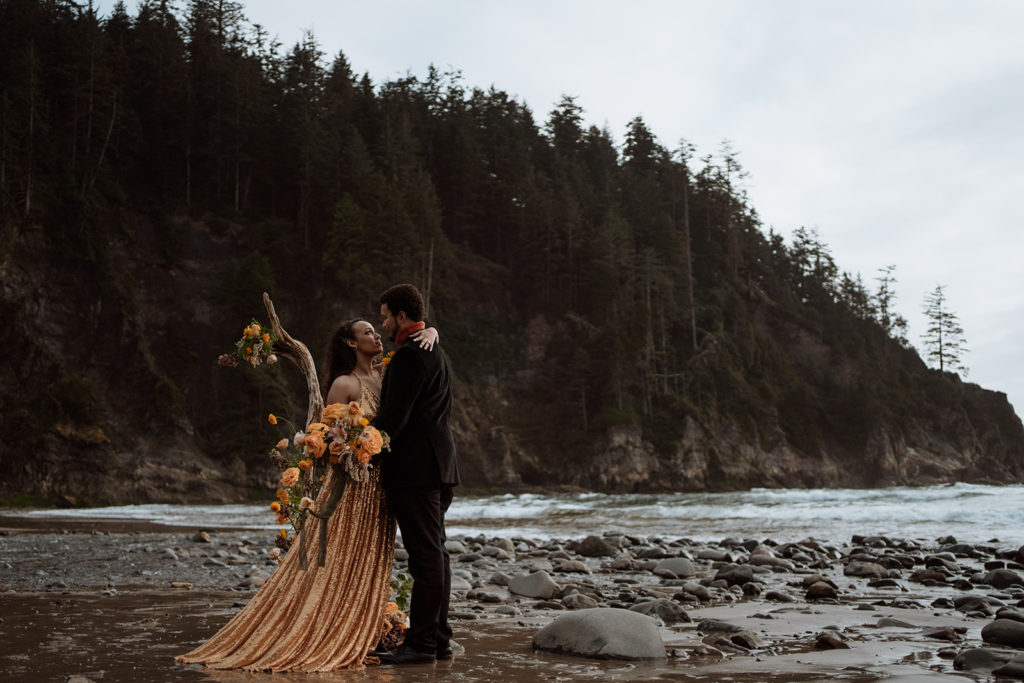 A bride in a gold dress stands with her new husband after eloping on the Oregon Coast.