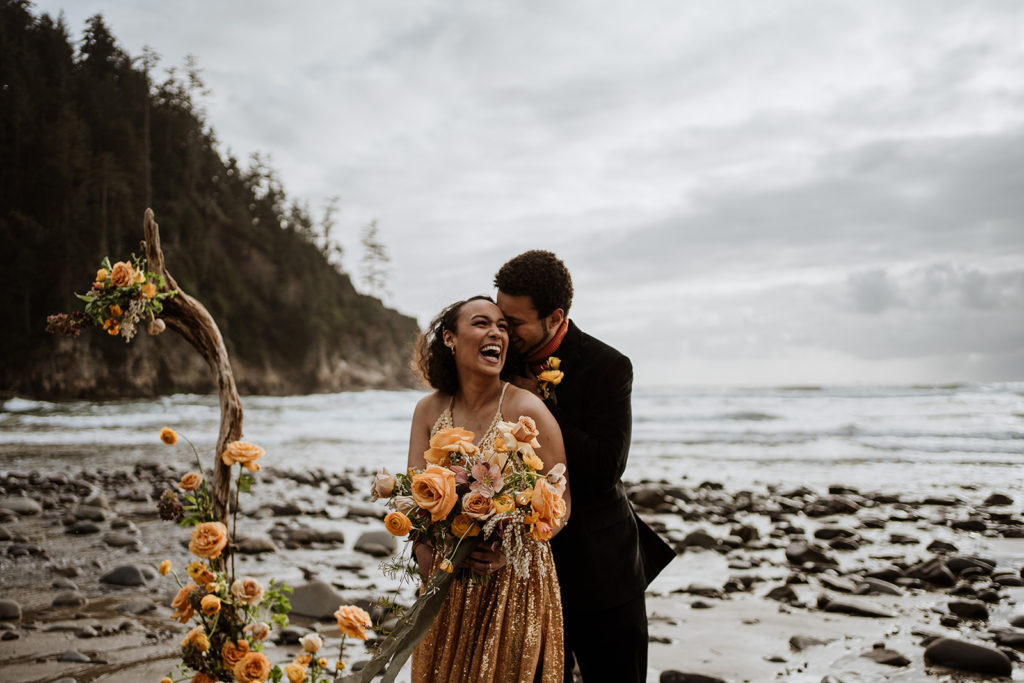 A bride in a gold dress is held by her husband after eloping on the beach in Oregon.