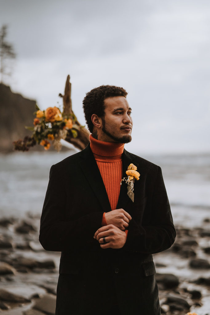 A groom stands on the beach overlooking the water.  He is wearing an orange turtle neck.  He has an orange rose boutonnière. 