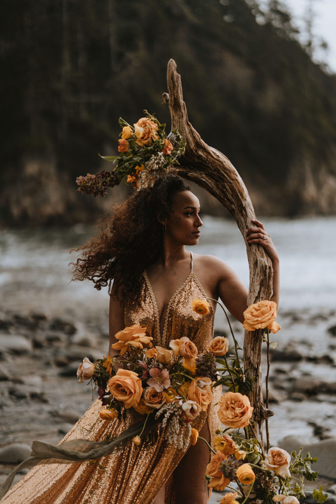 A bride stands next to driftwood surrounded by orange roses and wearing a gold and shimmering dress.