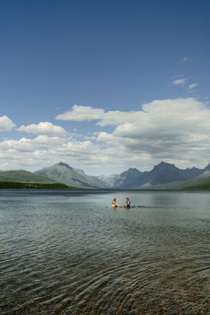 A couple sits on a paddle board on a lake in Glacier National Park in front of mountians.