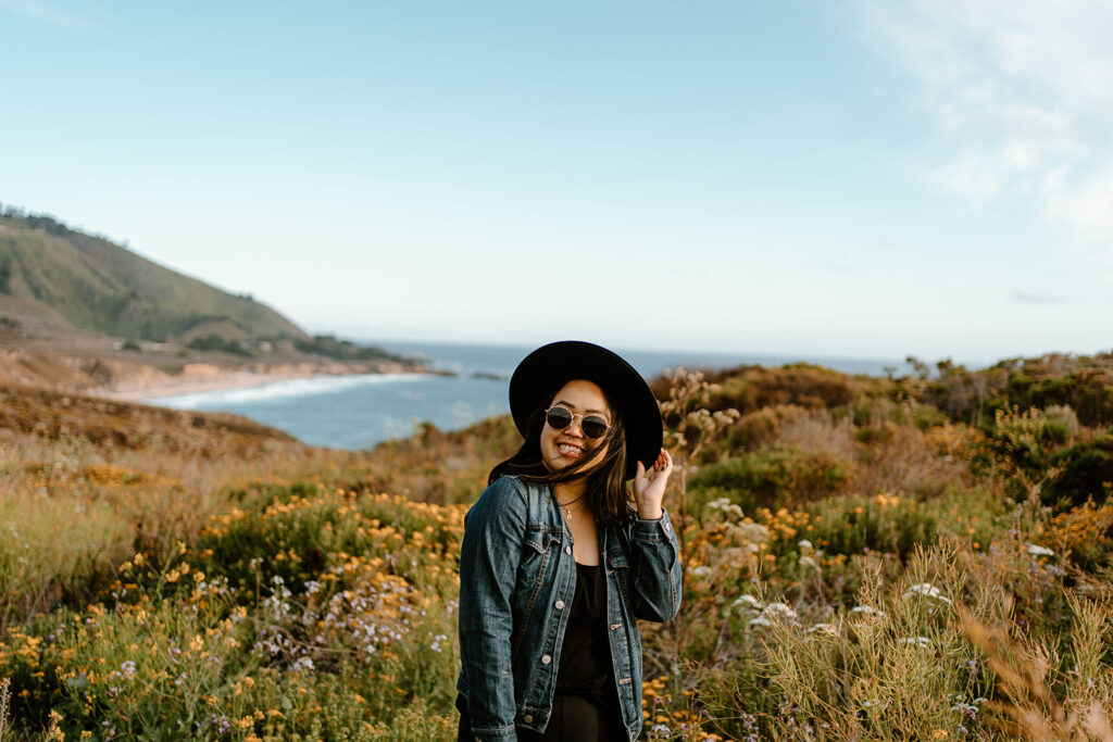 Photographer Kriztelle Halili stands in Big Sur national park smiling at the camera while standing in a field of flowers.  She's wearing a sun hat and standing in a field of flowers