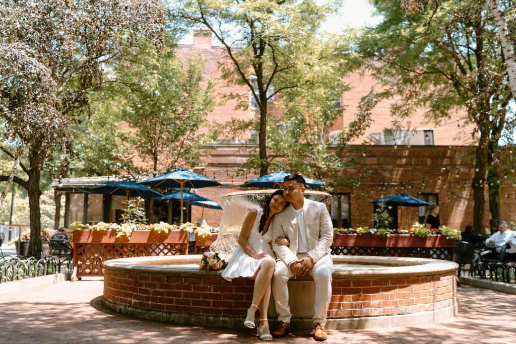 bride and groom sitting by a fountain with interlocking their arms on their elopement day