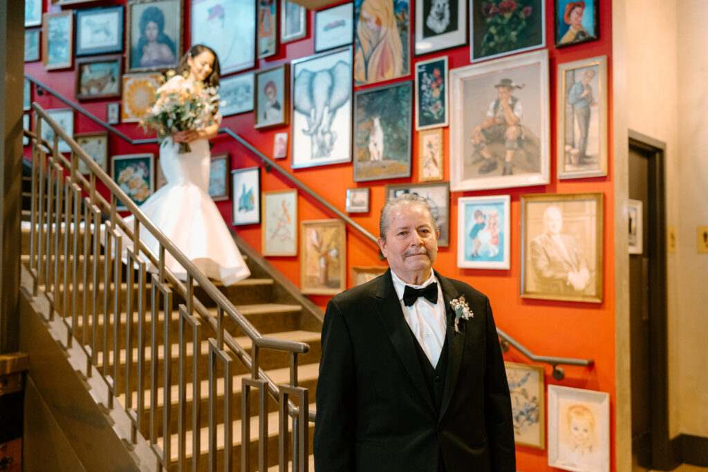 bride is coming down the stairs behind her father while he is facing the camera