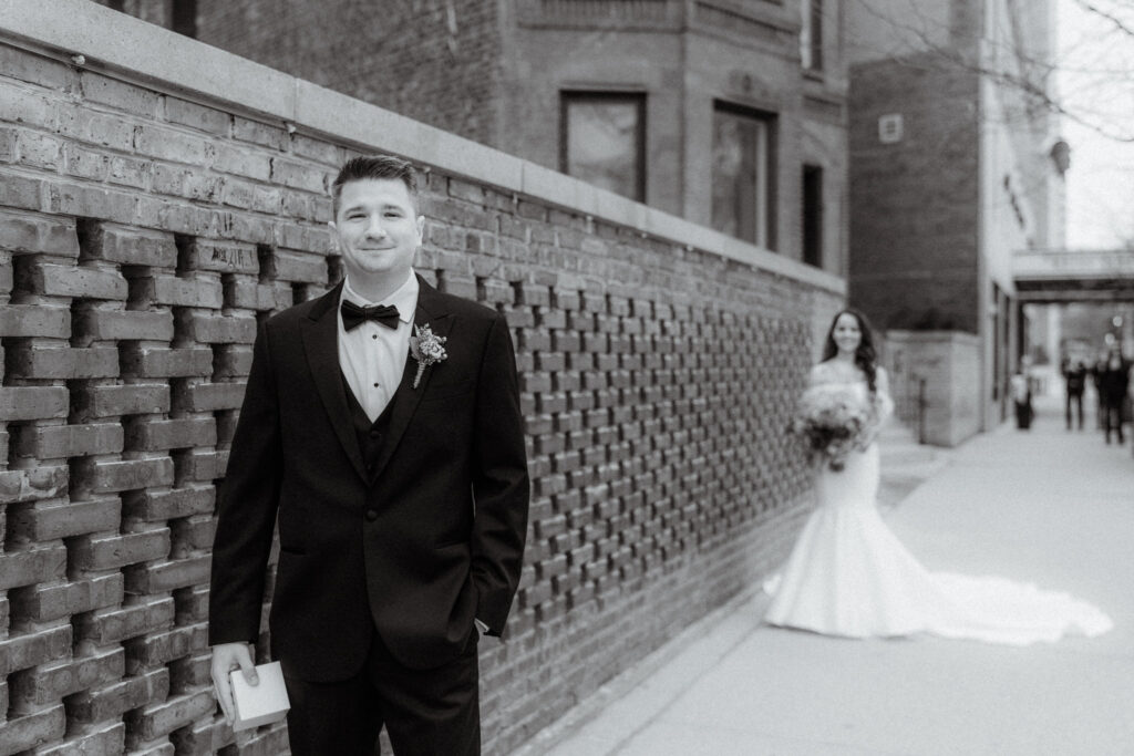 black and white photo of a groom facing the camera while his bride is standing behind him at a distance