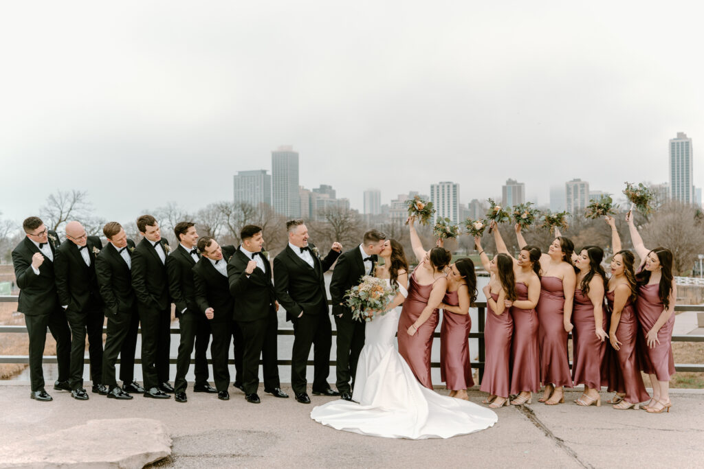 wedding party cheering on the bride and groom kissing with the Chicago skyline behind them