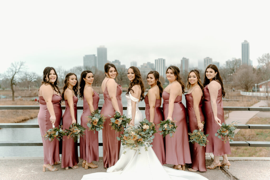 bride with her bridesmaids with the backs slightly facing the camera with one hand holding their bouquets down while looking at the camera; Chicago skyline as their background