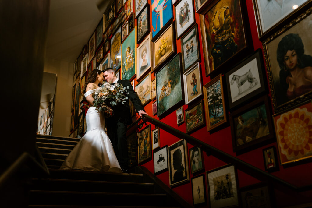 bride and groom with their foreheads together, spot light on them as they stand on stairway of red walls and filled with gallery walls