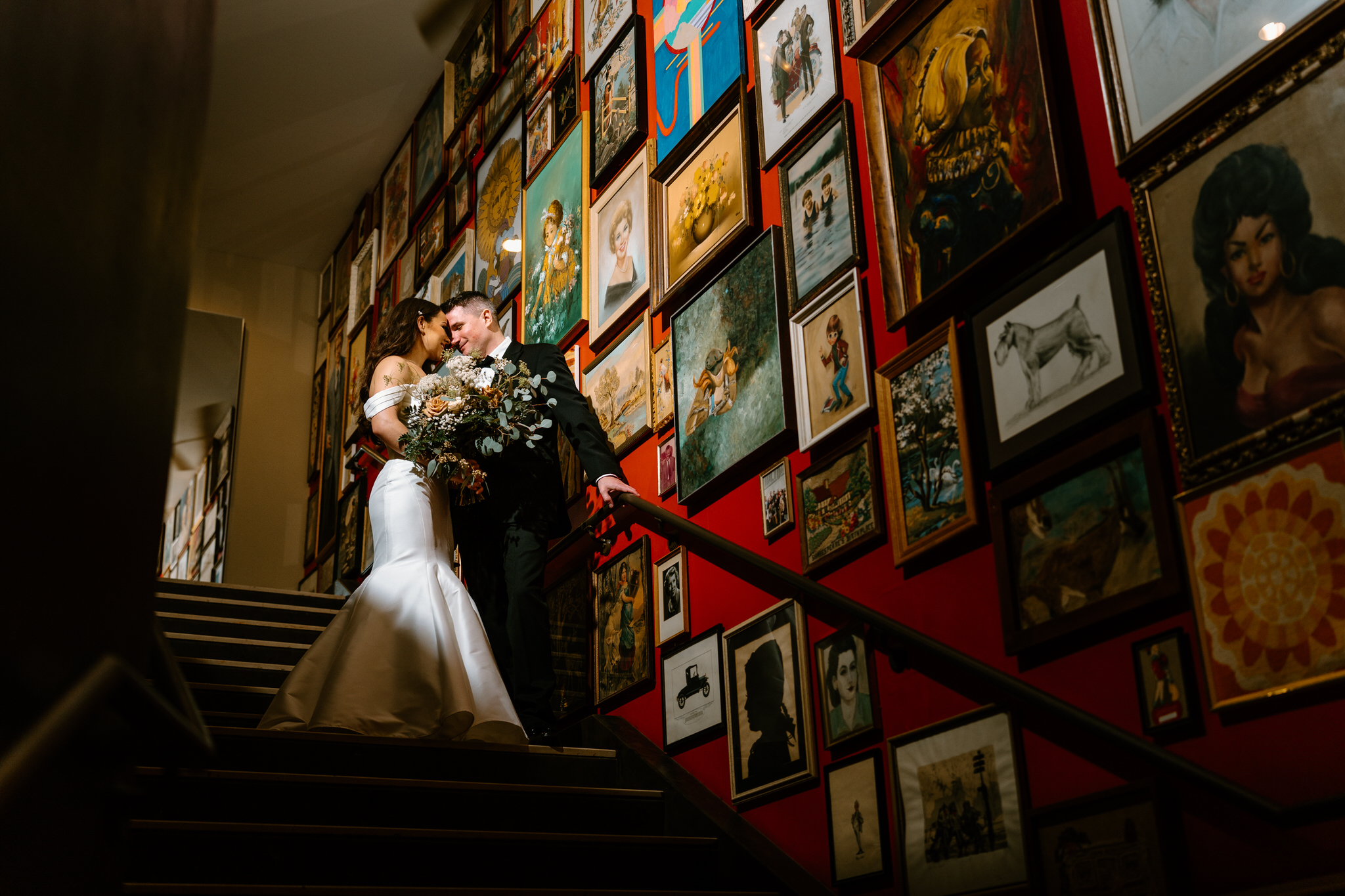 bride and groom with their foreheads together, spot light on them as they stand on stairway of red walls and filled with gallery walls