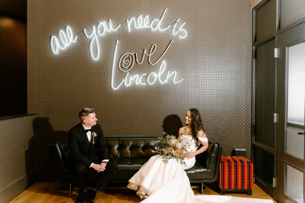 bride and groom sitting apart looking at each other; backdrop has neon lights saying "all you need is love lincoln"