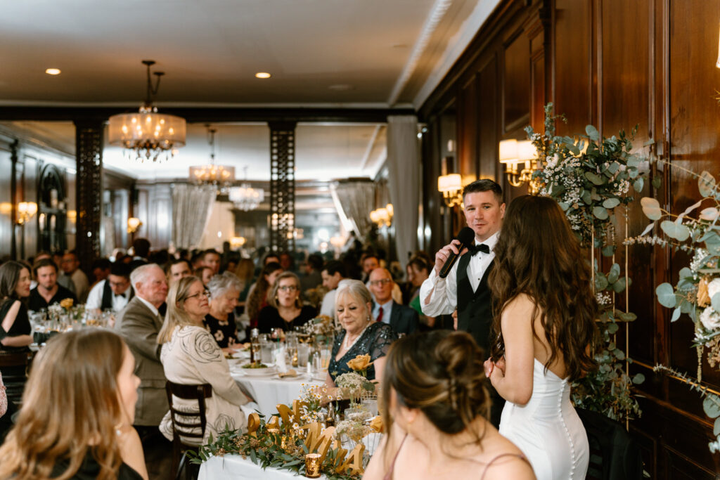 groom making a speech in front of their guests while looking at his wife